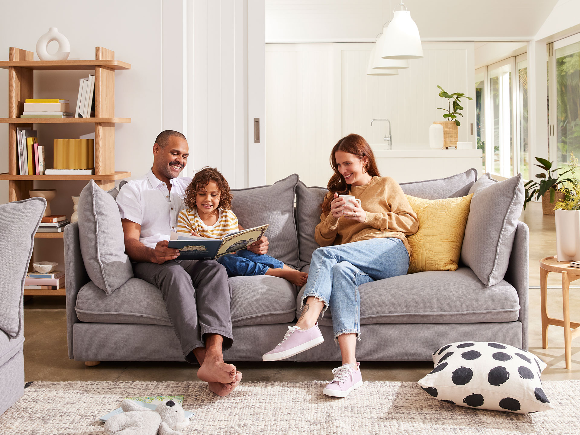 A father, mother and child sit on a three seater light grey sofa to read a book.