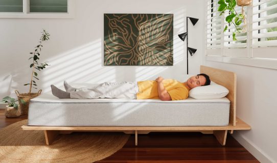 Man in yellow t-shirt lying down on a mattress trying to figure out if it’s the best mattress for him