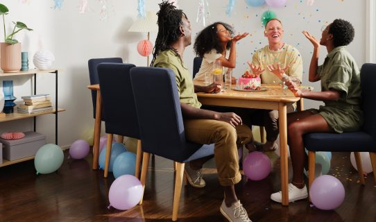 Four friends sit at a timber dining table with dark blue upholstered chairs, balloons and party streamers