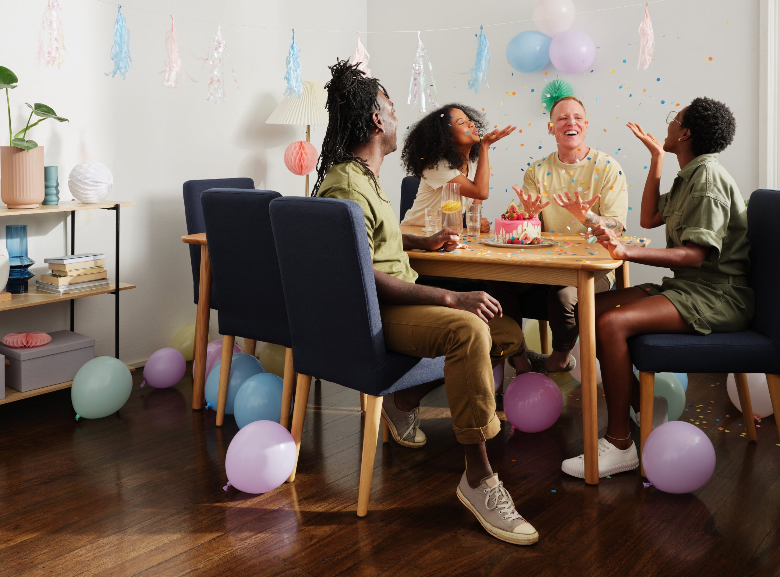 Four friends sit at a timber dining table with dark blue upholstered chairs, balloons and party streamers