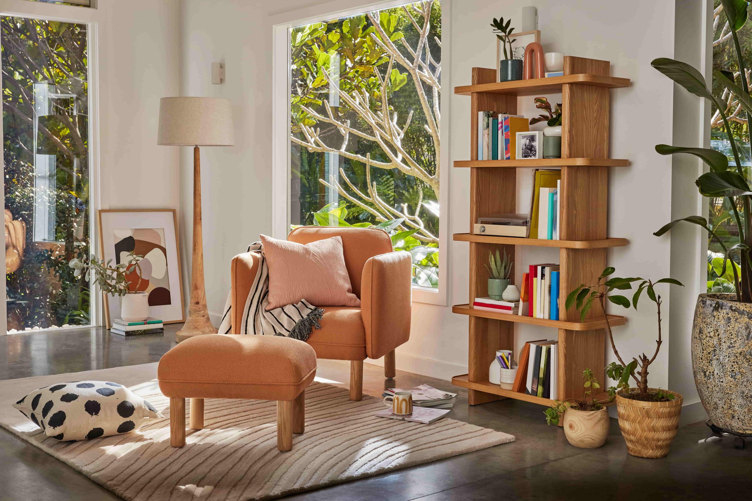An orange single seater armchair and ottoman with a pink cushion, black and white throw rug and timber bookshelf filled with books 
