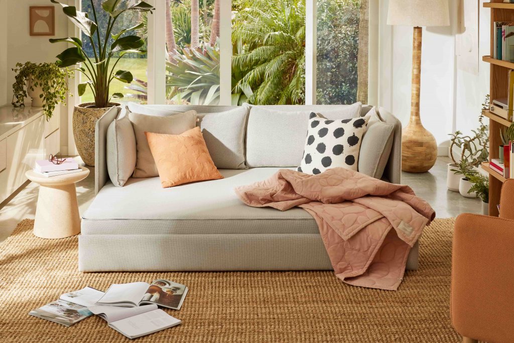 A light grey sofa bed with an orange and black and white cushions, pink throw and plants in the background