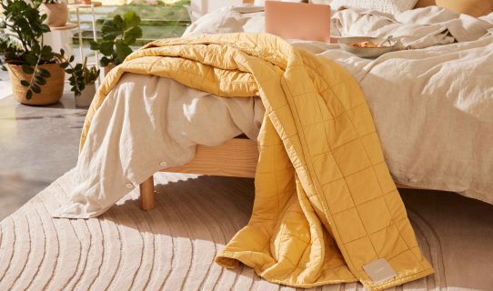 A textured cream rug with timber bed base on top, covered with linen bedding and a yellow throw