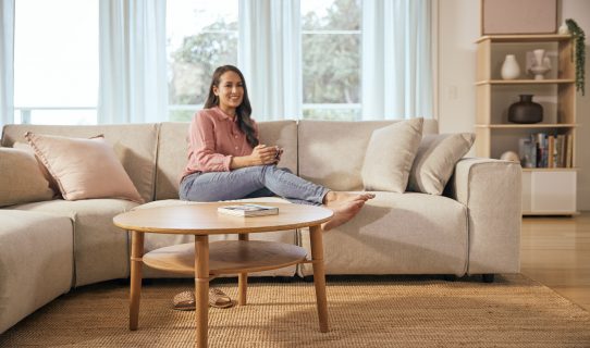 A smiling woman relaxes on her Koala sofa and behind her wood coffee table from Koala