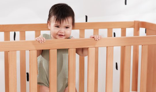 A baby stands up smiling in her cot, highlighting the importance of cot safety