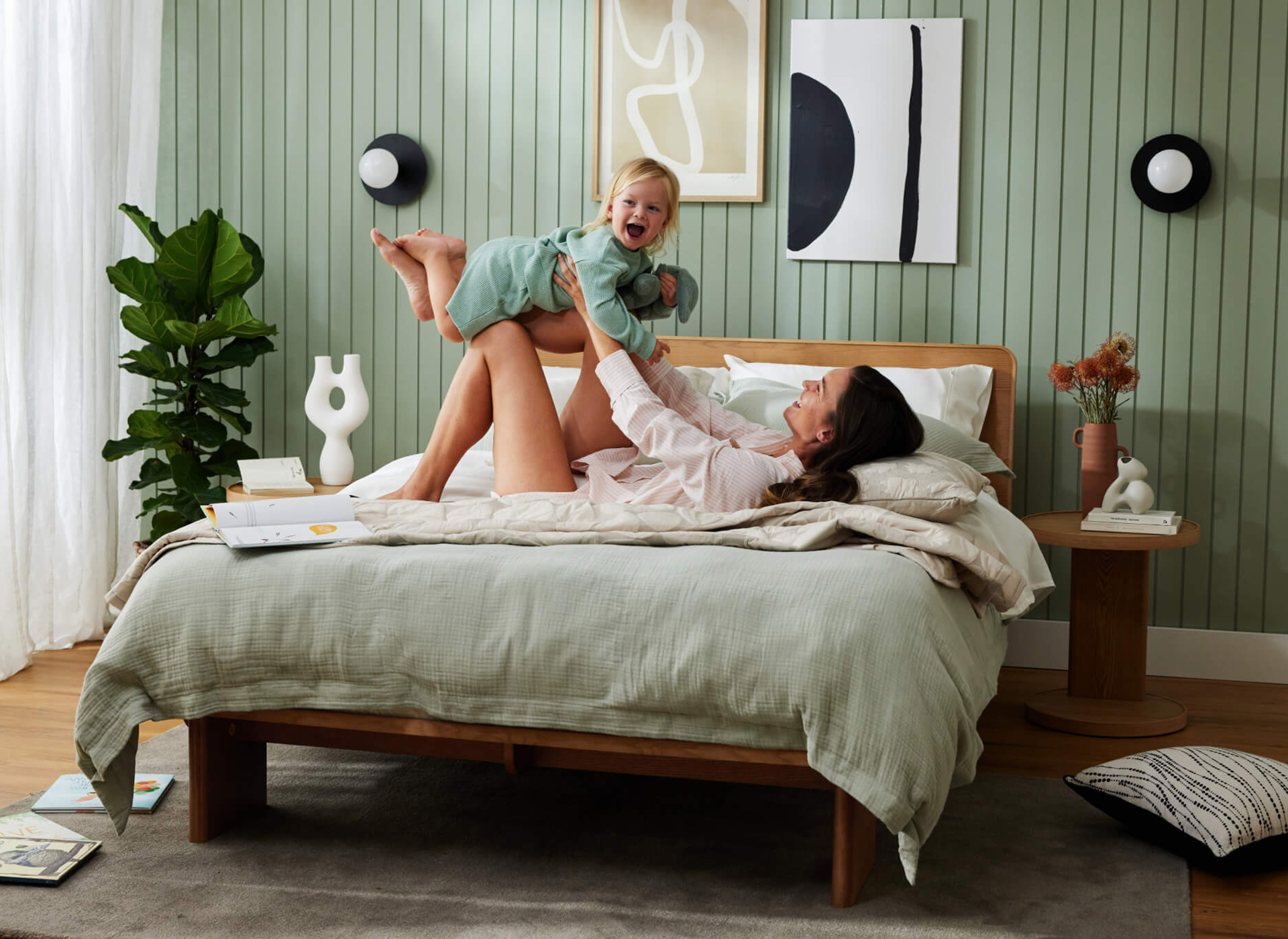 A mother and child play on their bed in a pretty room with green feature wall and timber bedroom furniture