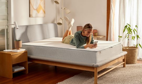 A woman lies on her Koala foam mattress and smiles down at her phone as she considers the advantages of a Koala mattress over a pocket springs mattress