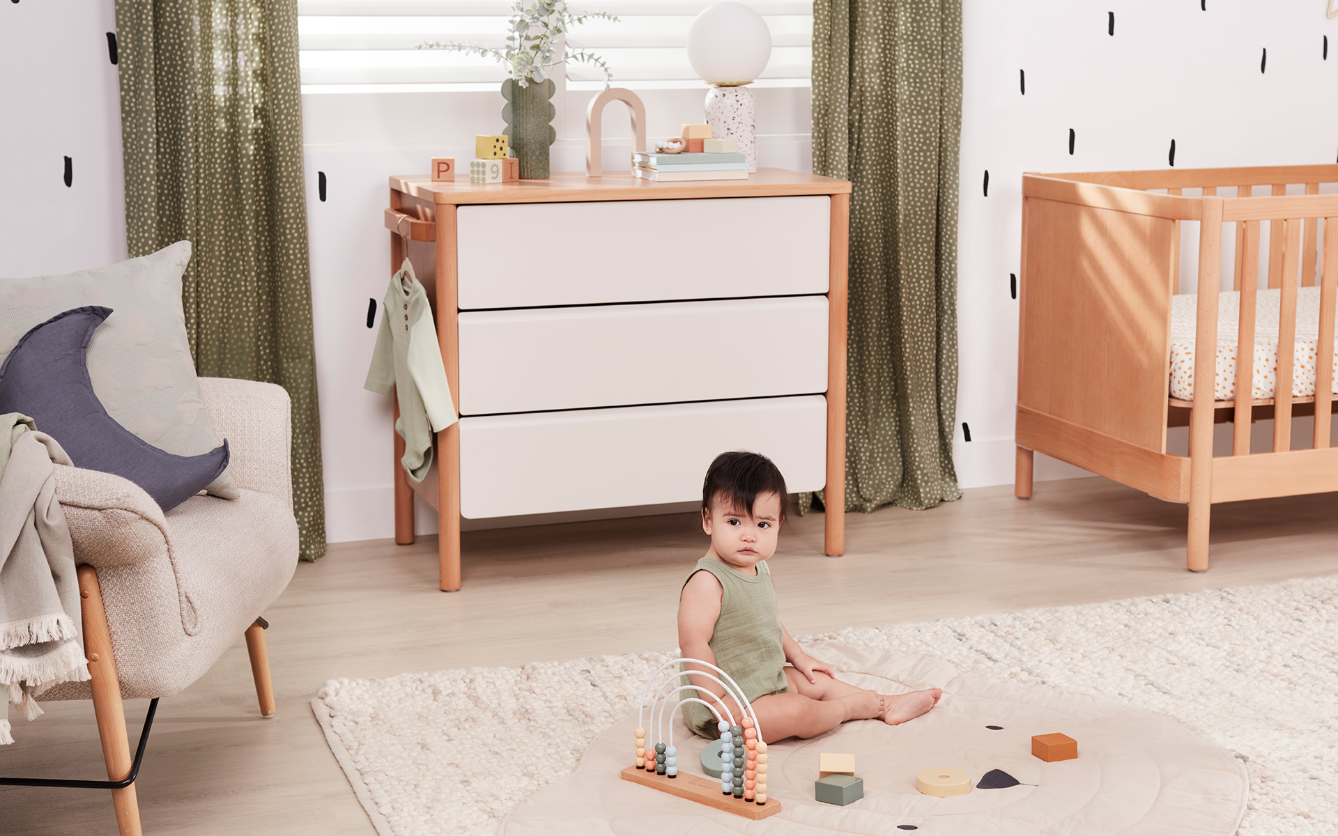 A baby sits on a rug in their room surrounded by kids furniture such as a cot and chest of drawers