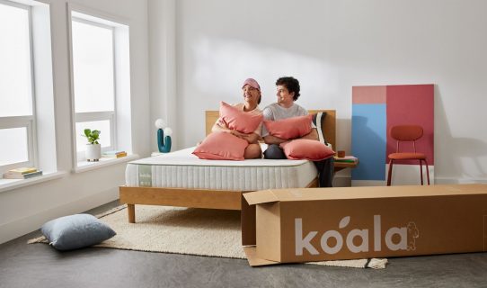 A couple sits on the Koala SE Mattress in their new house. The Koala bed box is featured in the image.